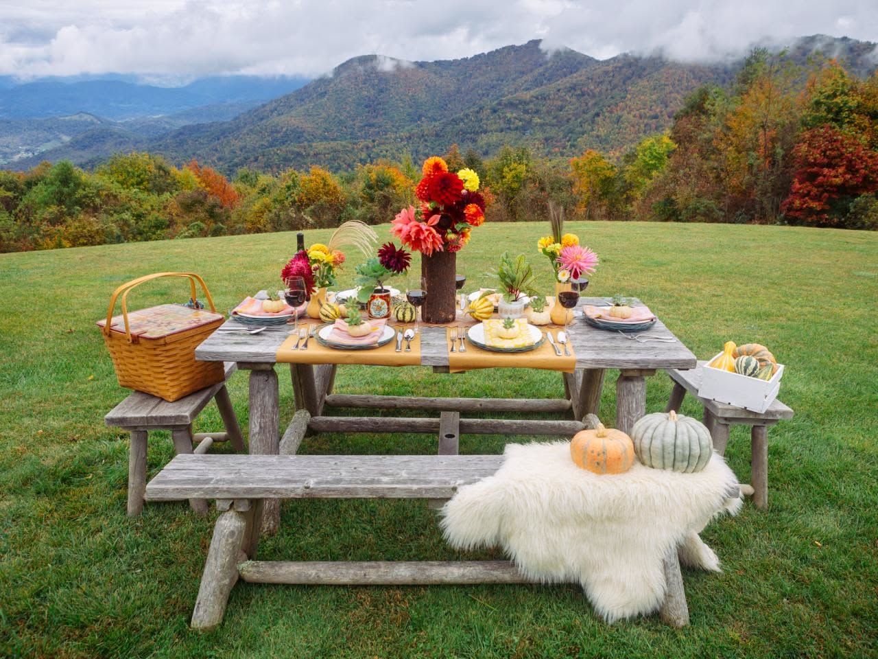 Rustic outdoor picnic table decorated with seasonal flowers and tableware set against a beautiful mountain landscape.